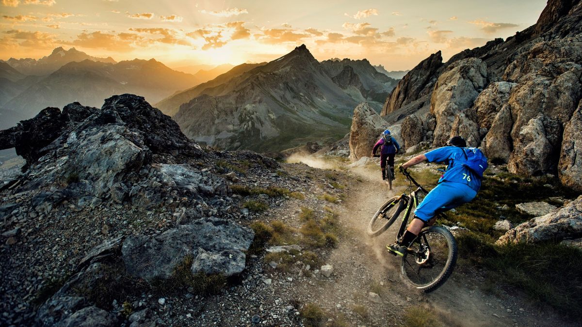 Two mountain bikers riding on a rocky trail at sunset, surrounded by rugged peaks and dramatic clouds.