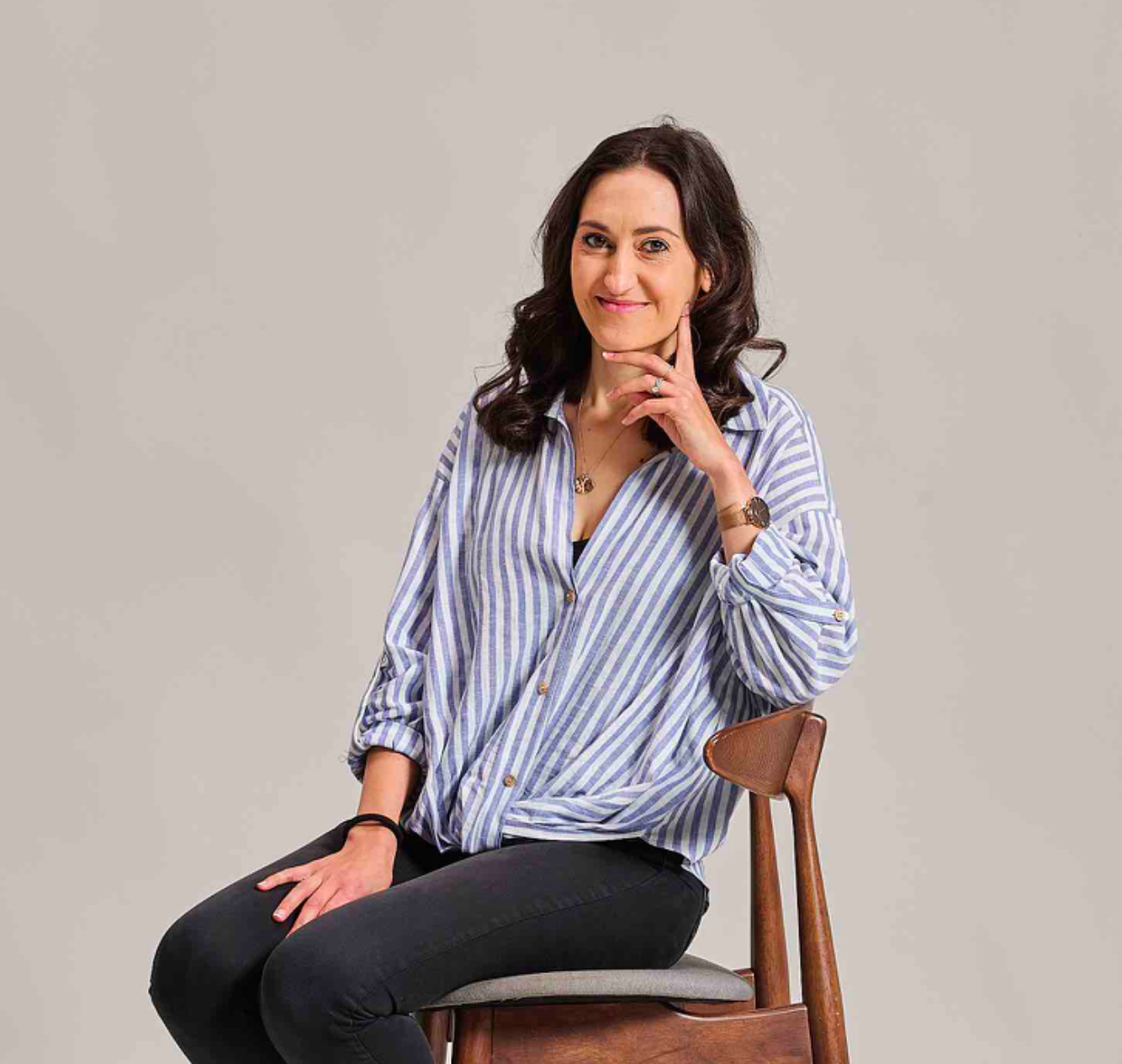 A woman in a striped shirt and black pants sits smiling on a wooden chair against a plain background.