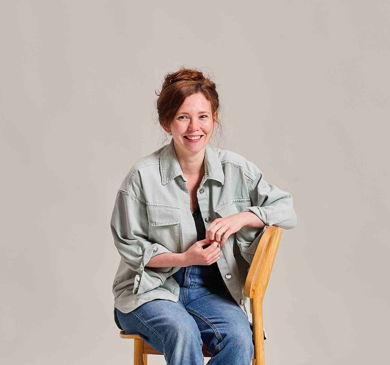 Smiling woman in casual shirt and jeans sits on a wooden chair against a plain background.