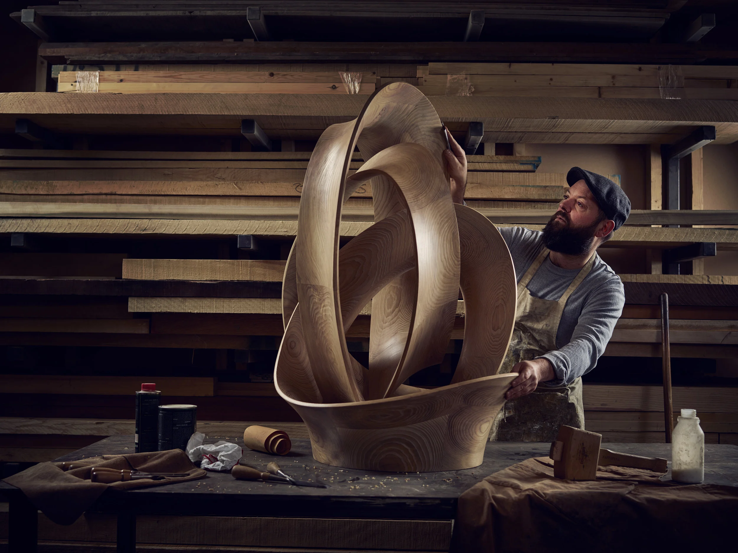 A bearded man shapes a large, abstract wooden sculpture in a workshop filled with wooden planks.